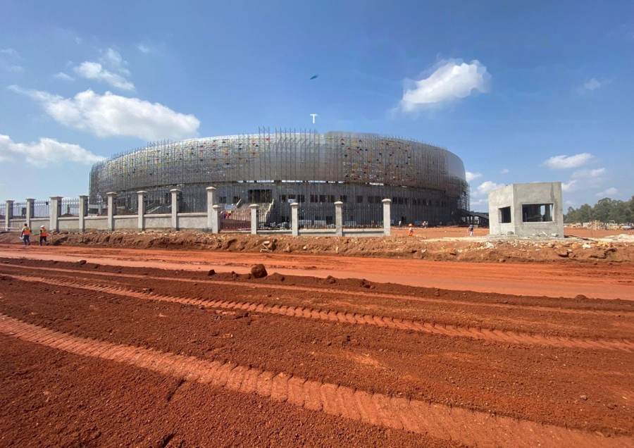 Access road formation and earthworks beside a large stadium structure under construction.